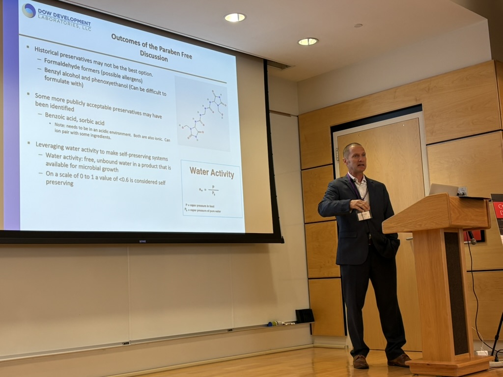A man in a suit gives a presentation at a podium with slides on "Outcomes of the Paraben Free Discussion" displayed on a large screen in a classroom or lecture hall.