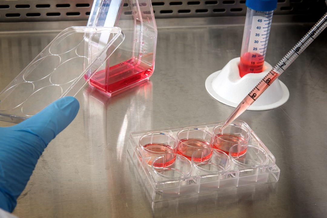 A gloved hand uses a pipette to transfer pink liquid into a six-well cell culture plate on a lab bench, illustrating the precision required in process development and pharmaceutical analytical method validation. Other laboratory containers are nearby.
