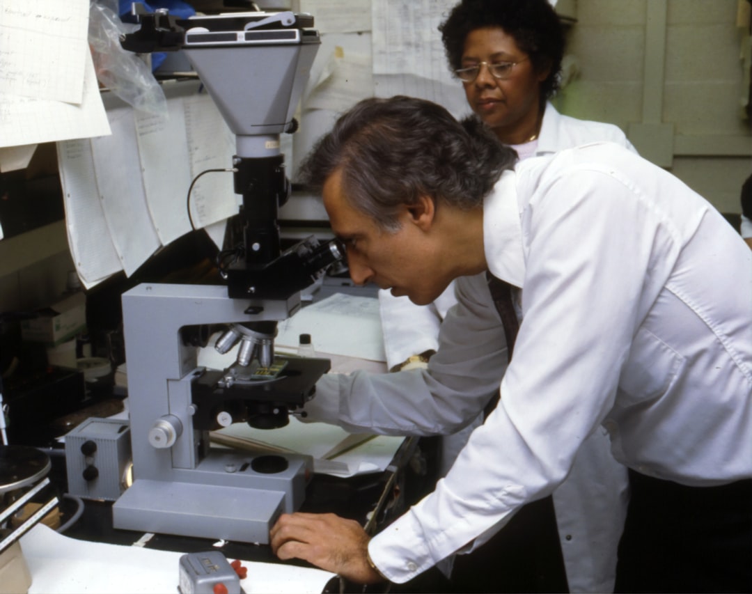 A man examines a specimen under a microscope while a woman observes in a laboratory filled with equipment and papers, engaged in analytical method development for ophthalmic product research.