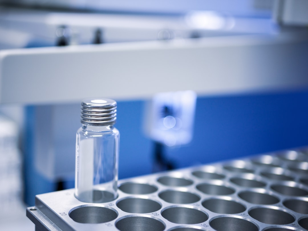 A small glass vial with a metal cap sits in a metal rack with circular slots, used for ICH stability testing in semi-solid dosage form development, set against a laboratory background with blurred equipment.